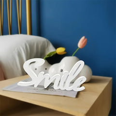 Decorative sign with 'Smile' on a table with flowers and pillows in the background.