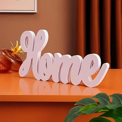 Decorative word 'Home' on a wooden surface with a plant and framed picture in the background.