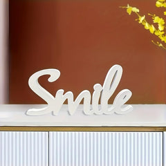 White decorative sign with 'Smile' on a shelf against a brown wall with yellow flowers.