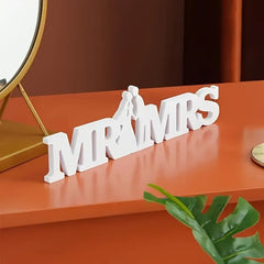 Decorative sign with 'MR & MRS' on an orange surface with a mirror and plant in the background.