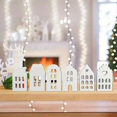 Decorative white houses on a wooden surface with a festive background featuring lights and a fireplace.