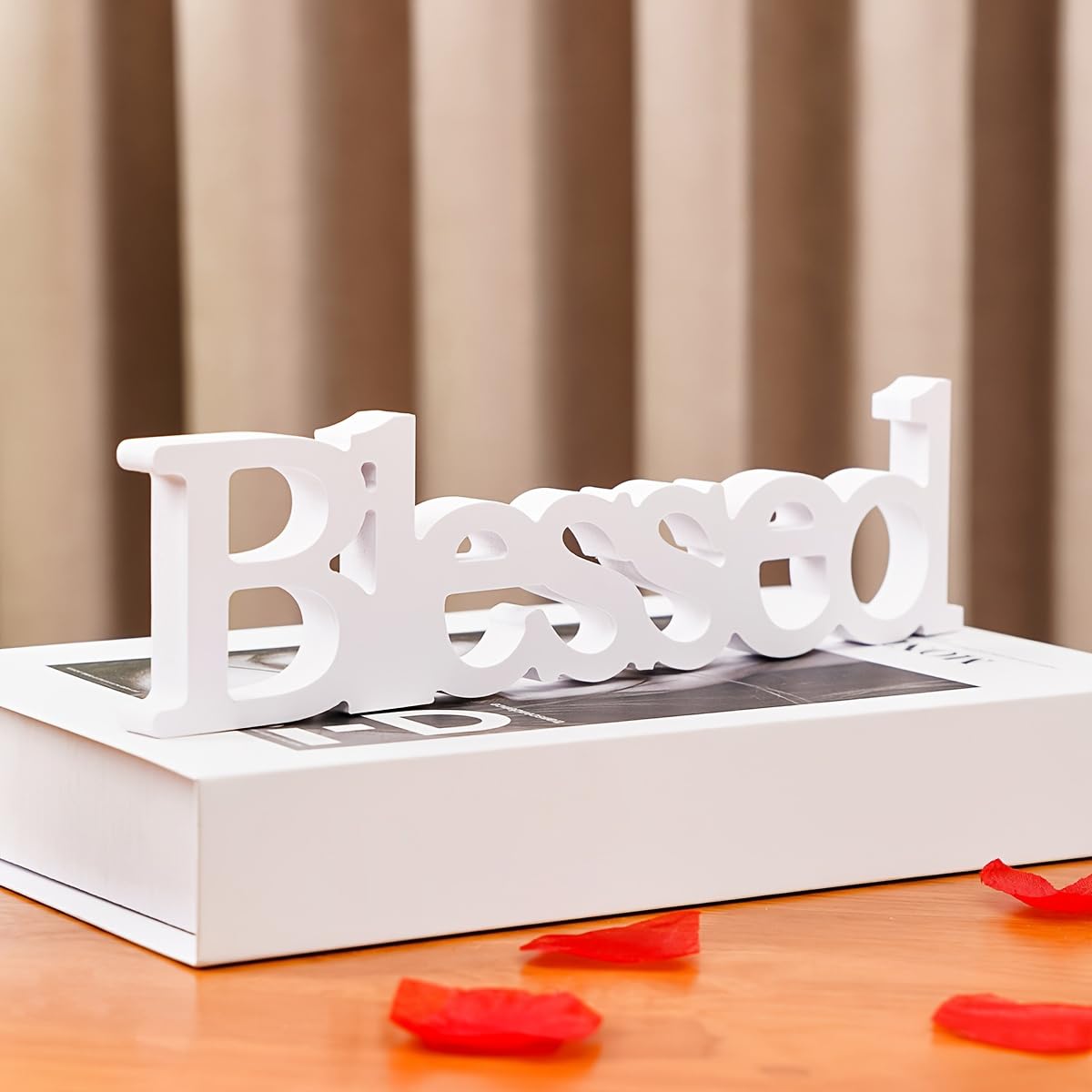 White decorative word 'Blessed' on a book with red rose petals on a wooden surface.