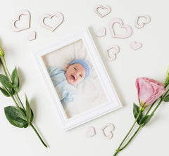 White photo frame with a baby in a blue outfit, surrounded by heart-shaped decorations and pink flowers on a white background.