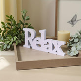 White 'Relax' sign on a wooden surface with plants and a candle in the background