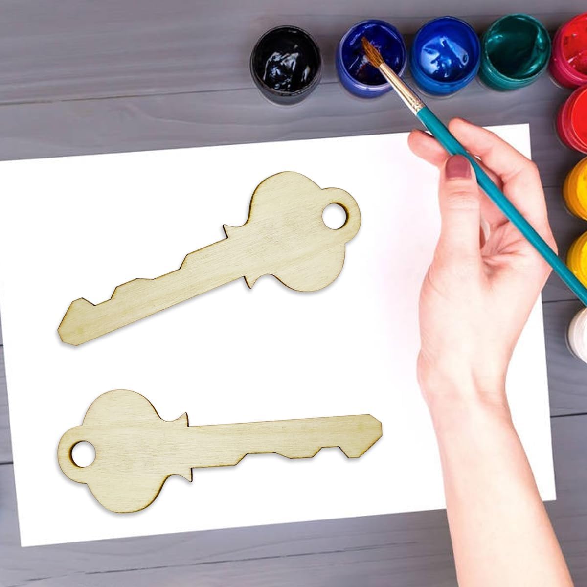 Person painting wooden key shapes with a brush on a white sheet of paper.