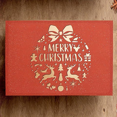 Red Christmas card with white 'Merry Christmas' text and festive illustrations on a wooden surface.