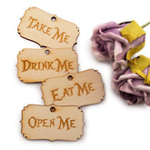 Wooden tags with text 'Take Me', 'Drink Me', 'Eat Me', and 'Open Me' on a white background with flowers.