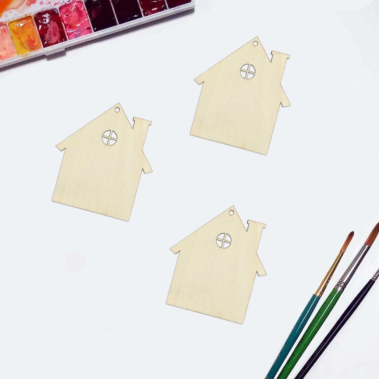 Three house-shaped wooden ornaments with paintbrushes and watercolors on a white background