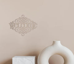Decorative 'welcome' wall art on a beige wall with a white vase and box below.