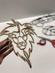 Hand holding a cut-out paper design of a lion's head with red accents on a white surface.
