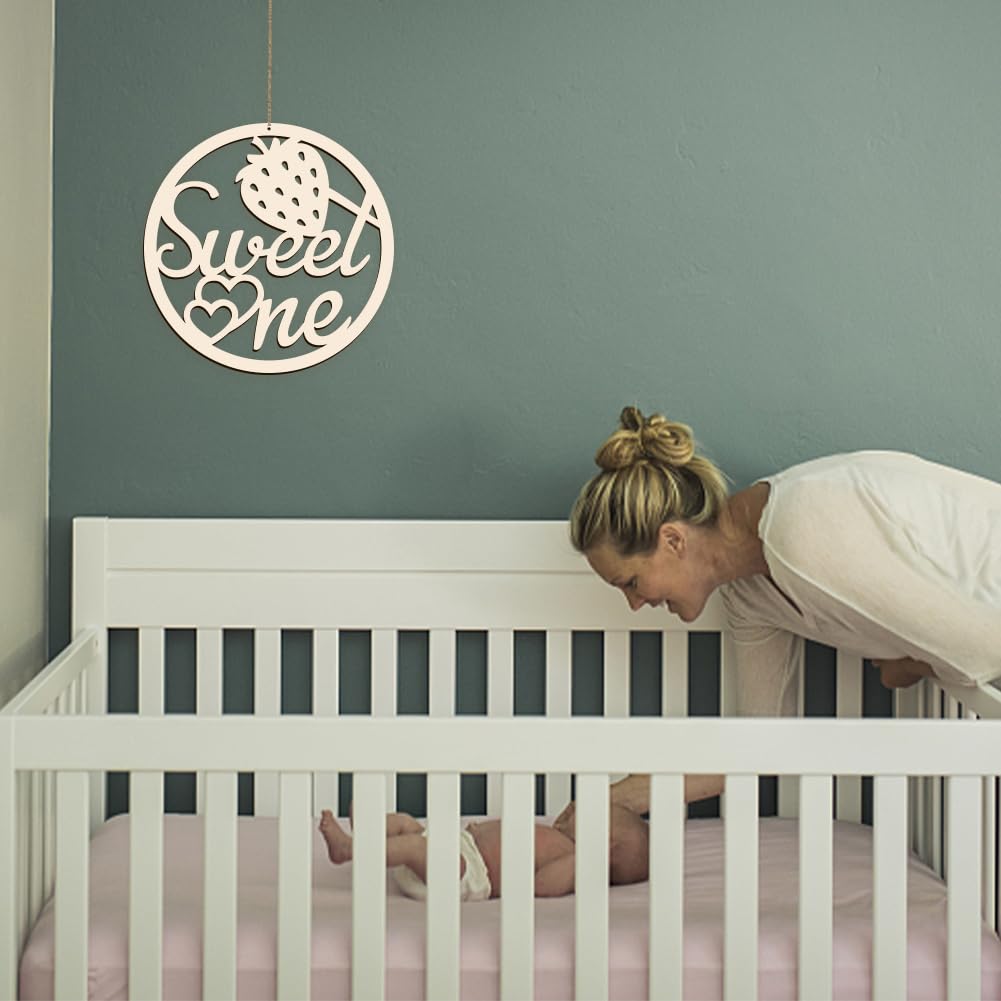 Woman leaning over a crib with a 'Sweet One' sign on the wall.