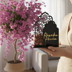 Decorative lantern with 'Ramadan Kareem' text held by a person in front of pink flowers.