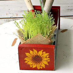 Red wooden planter with sunflower design containing green plants and gardening tools on a white surface.