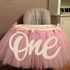 High chair with pink tulle skirt and 'One' decoration against a neutral wall.