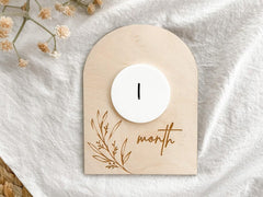 Wooden baby milestone card with a white candle and '1 month' text on a white fabric background.