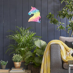 Colorful unicorn wall art on a dark wooden panel wall with plants and a gray chair.