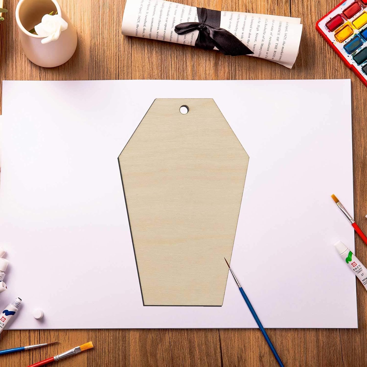Beige paper cutout of a coffin shape on a white sheet with art supplies on a wooden table.
