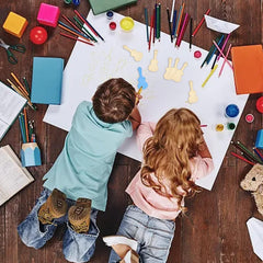 Two children sitting at a table with art supplies, drawing on a large sheet of paper.