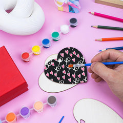 Person painting a heart-shaped craft item with various art supplies on a pink background