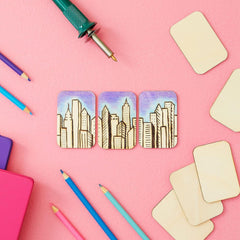 Three wooden coasters with cityscape design on a pink background with art supplies.