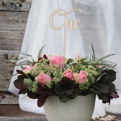 Floral arrangement with a 'One' cake topper on a wooden surface.