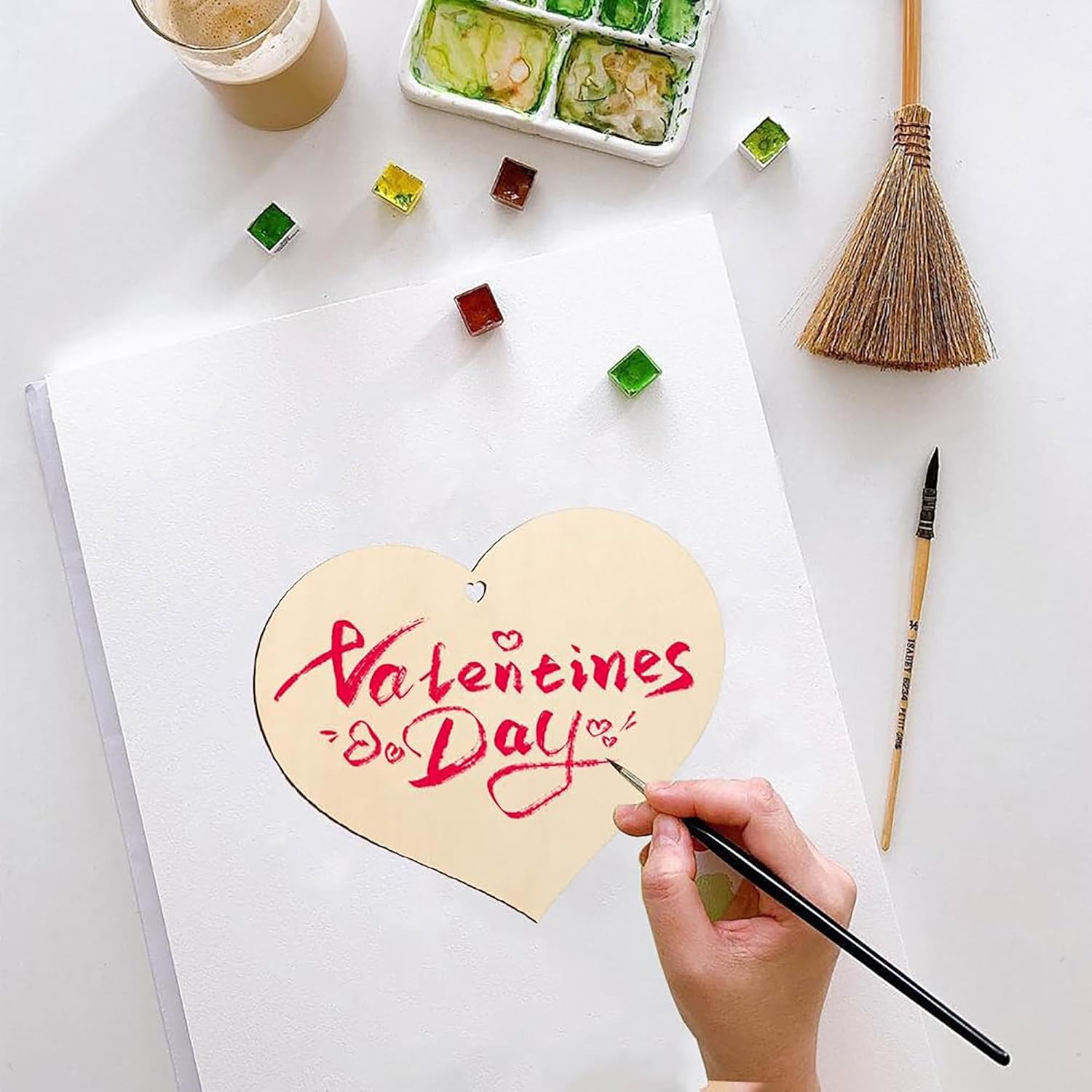 Heart-shaped card with 'Valentine's Day' writing being painted on a white surface with art supplies.