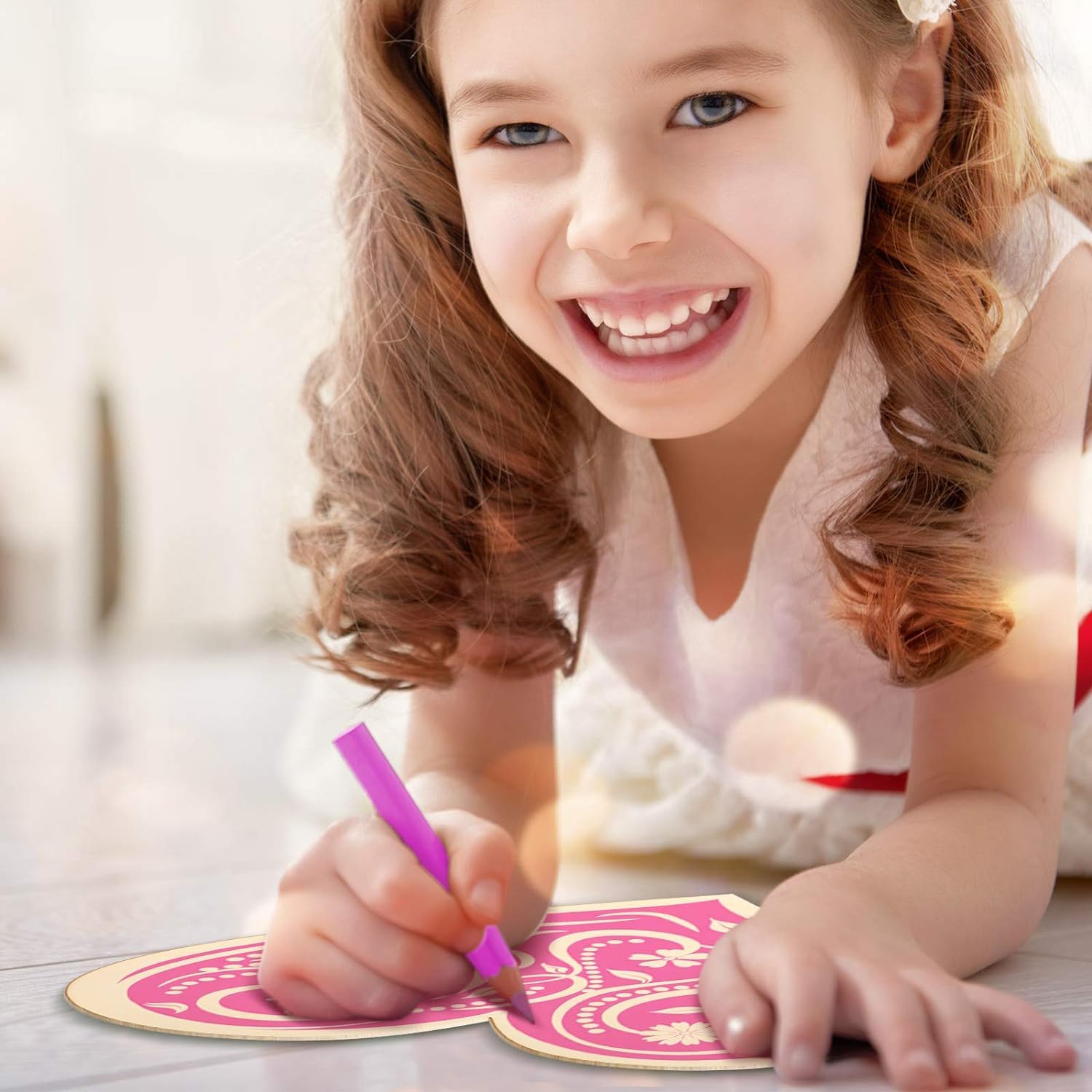 Young girl smiling and drawing with a pink crayon on a white surface.