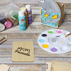 Painting supplies including paint bottles, a palette with colors, and a wooden coaster with 'Mom Pamela' engraved on a wooden surface.