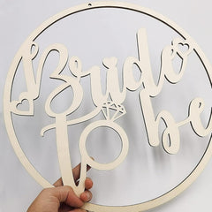 Wooden decorative sign with 'Bride & Be' text and a ring design, held against a light gray background.
