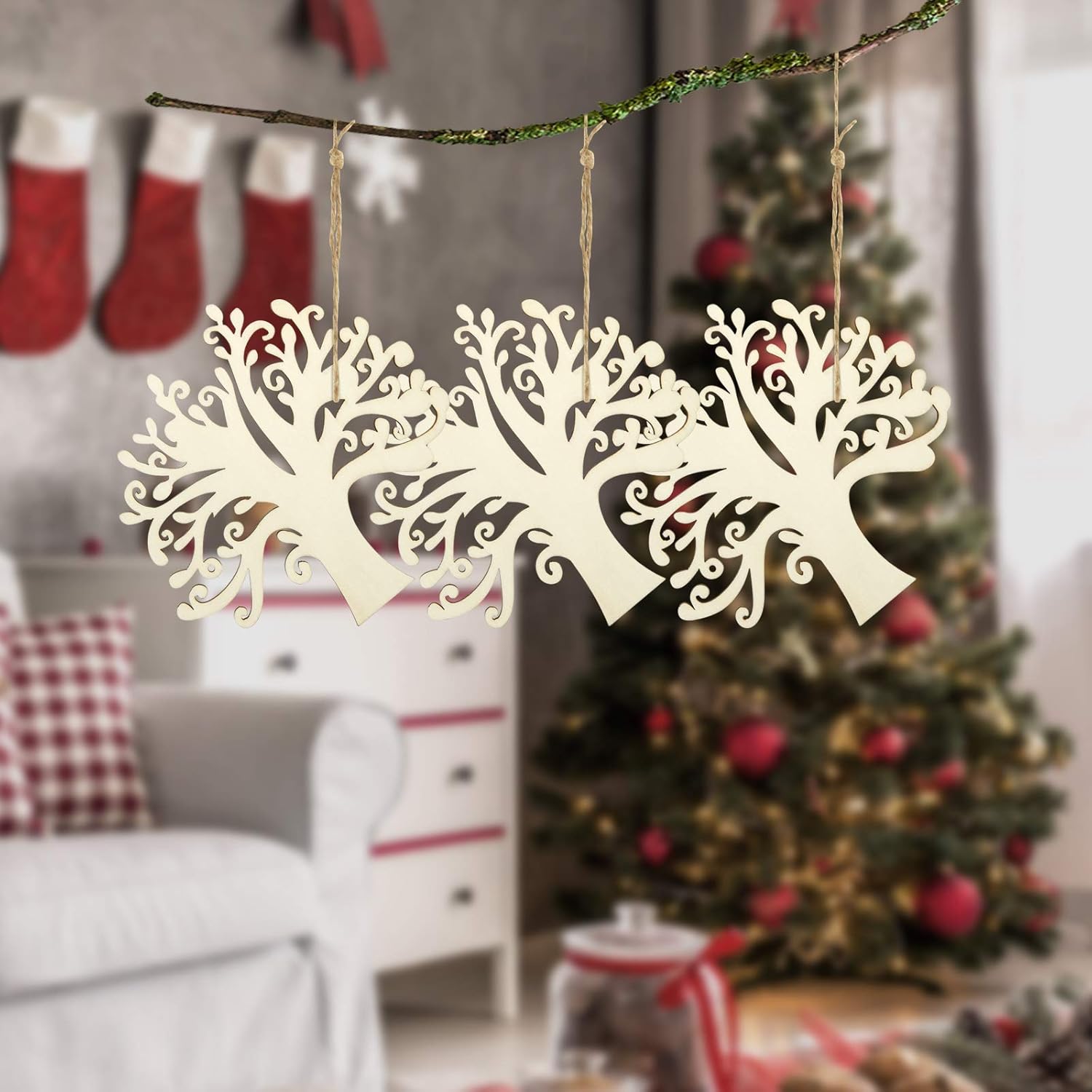 Decorative tree-shaped ornaments hanging on a branch with a Christmas tree and stockings in the background.