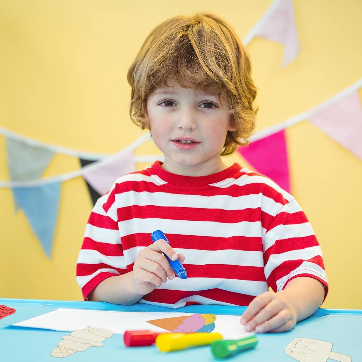 Child in a red and white striped shirt holding a blue marker, sitting at a table with colorful markers and paper.