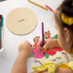 Child coloring a floral design on paper with a wooden circle and pencils on a table.
