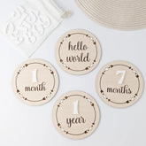 Wooden milestone discs for baby's first year with text like 'hello world', '1 month', '7 months', and '1 year' on a light background.