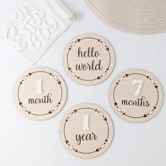Wooden milestone discs for baby's first year with text like 'hello world', '1 month', '7 months', and '1 year' on a light background.