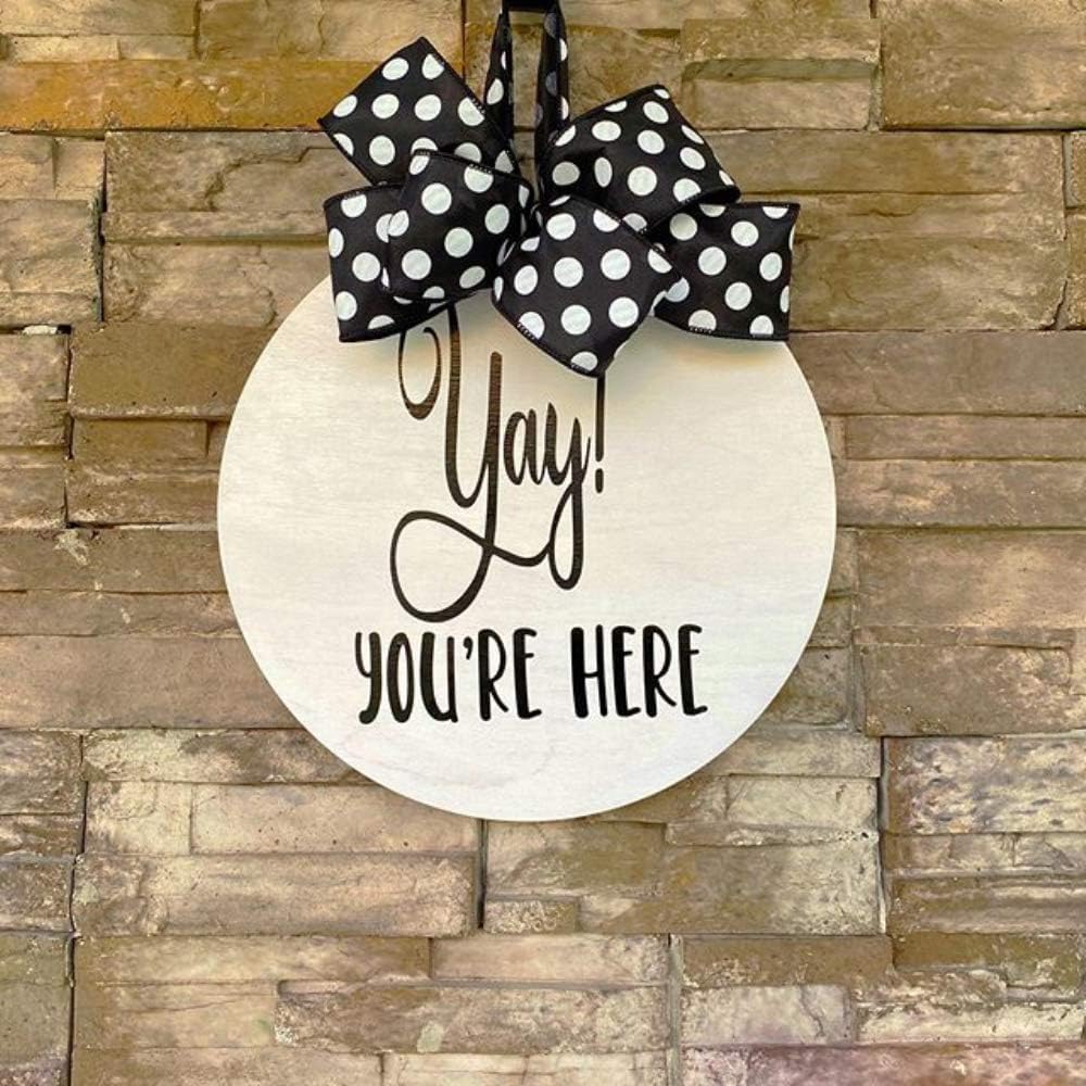 Round wooden sign with 'Yay! You're Here' text and black polka dot bow on a stone wall background