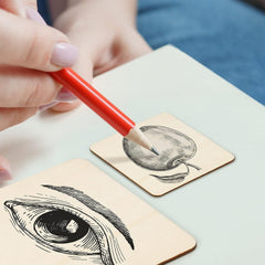 Person holding a red pencil over two wooden blocks with black ink illustrations of an eye and a peach.