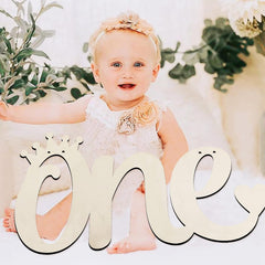 Baby in a white outfit with floral headband sitting behind large letter 'one' decorations.