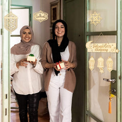 Two women holding small gift boxes in front of a decorated door with 'Ramadan Kareem' signs.