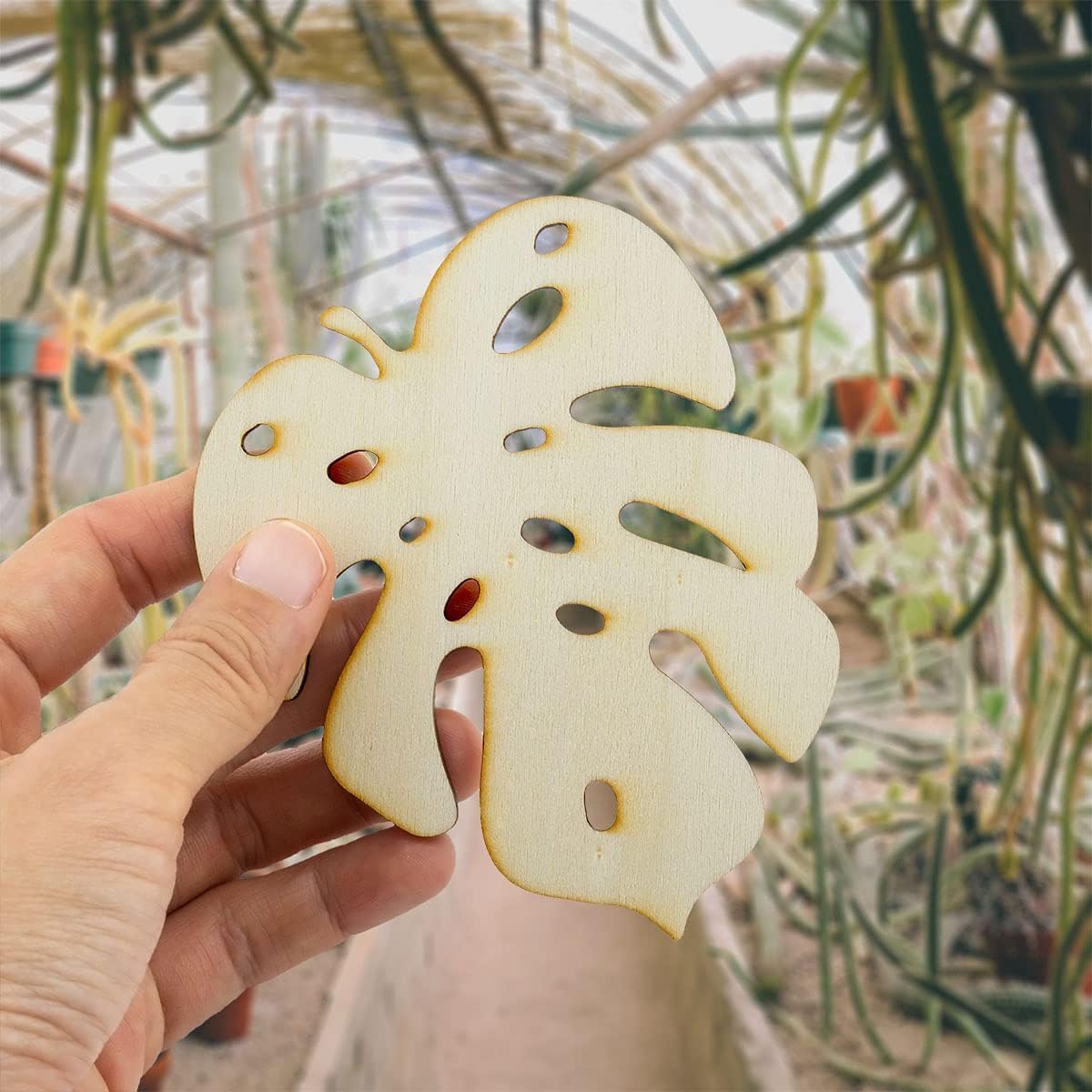 Hand holding a leaf-shaped object with a plant-filled greenhouse in the background