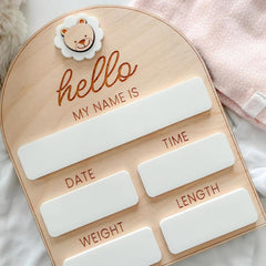 Wooden baby milestone board with white labels and a teddy bear charm on a soft pink background.