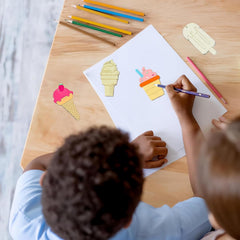 Two children drawing ice cream cones on a piece of paper with colored pencils on a wooden table.