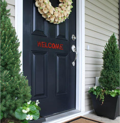 Black front door with a wreath and 'WELCOME' sign, flanked by potted plants.