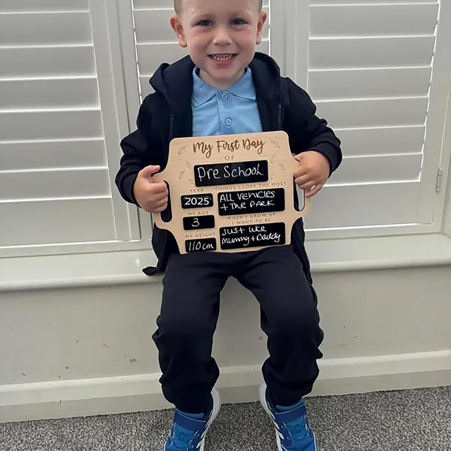 Child holding a sign for their first day of preschool in front of white blinds.