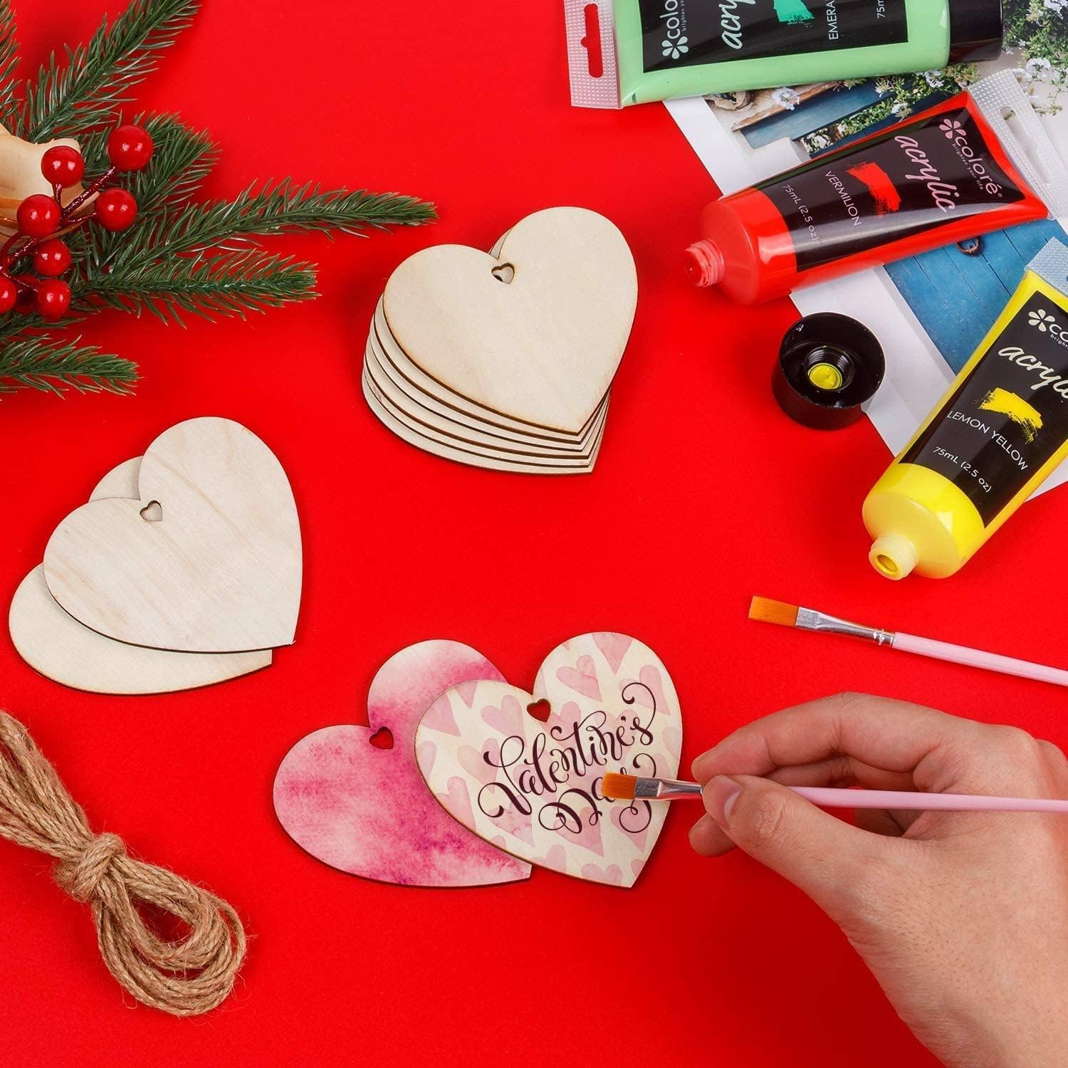 Hand painting a wooden heart ornament with 'Valentine's' on a red surface with art supplies.