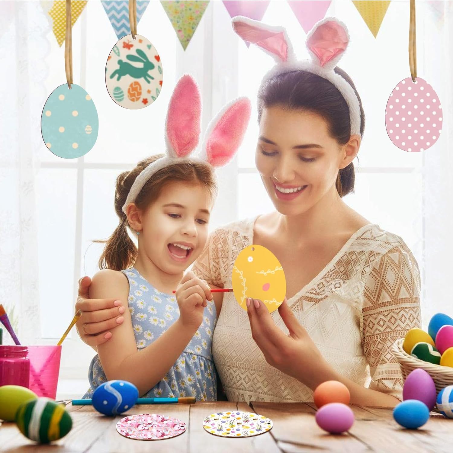 Woman and child wearing bunny ears, painting Easter eggs with colorful decorations in the background.