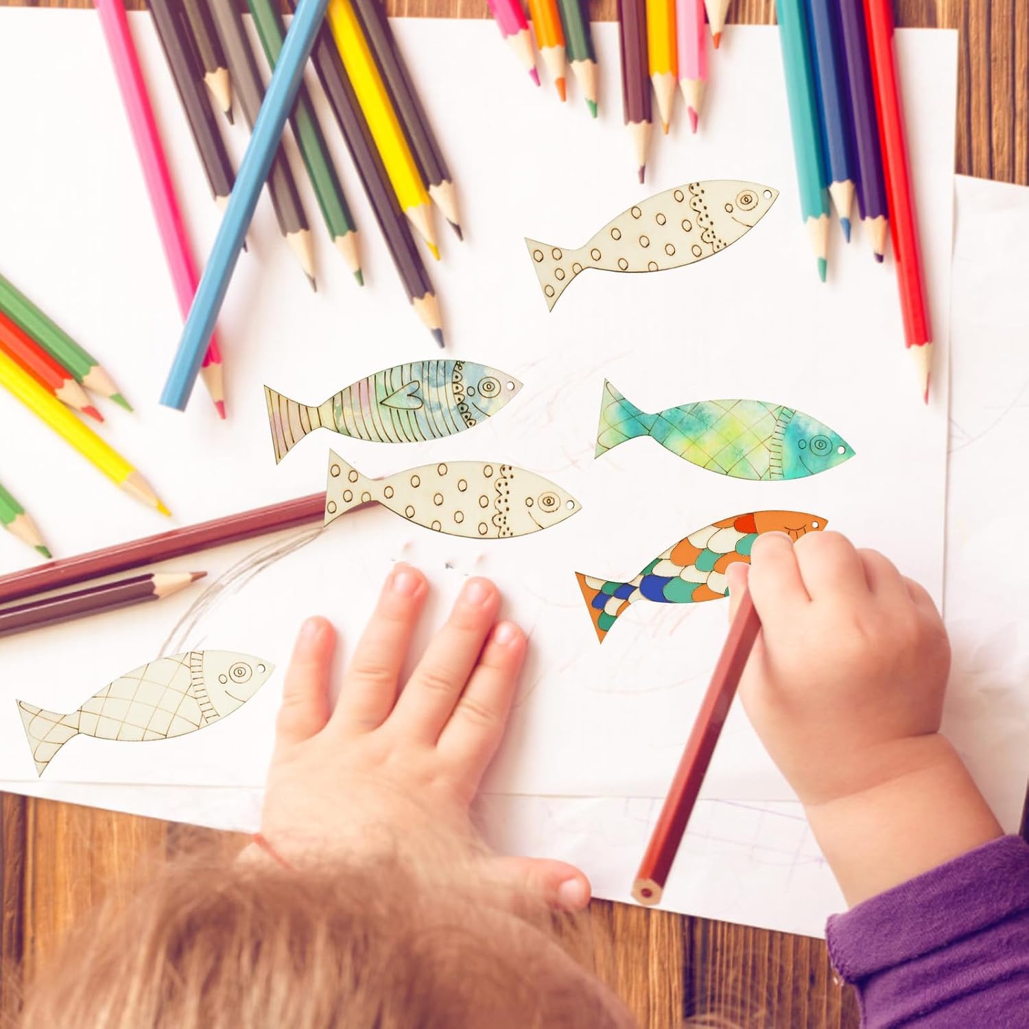Children coloring fish illustrations with colored pencils on a wooden table.