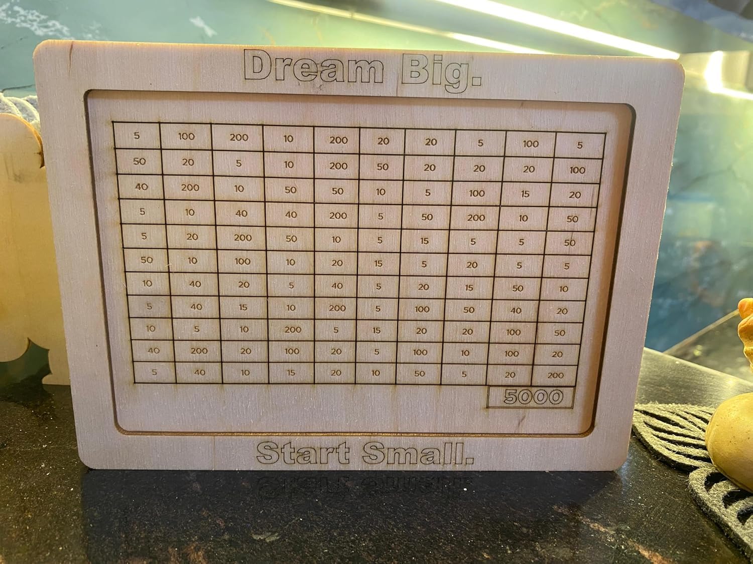 Wooden plaque with a grid and motivational text 'Dream Big. Start Small.' on a reflective surface.