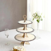 Three-tiered decorative stand on a table with a glass, flowers, and cupcakes.