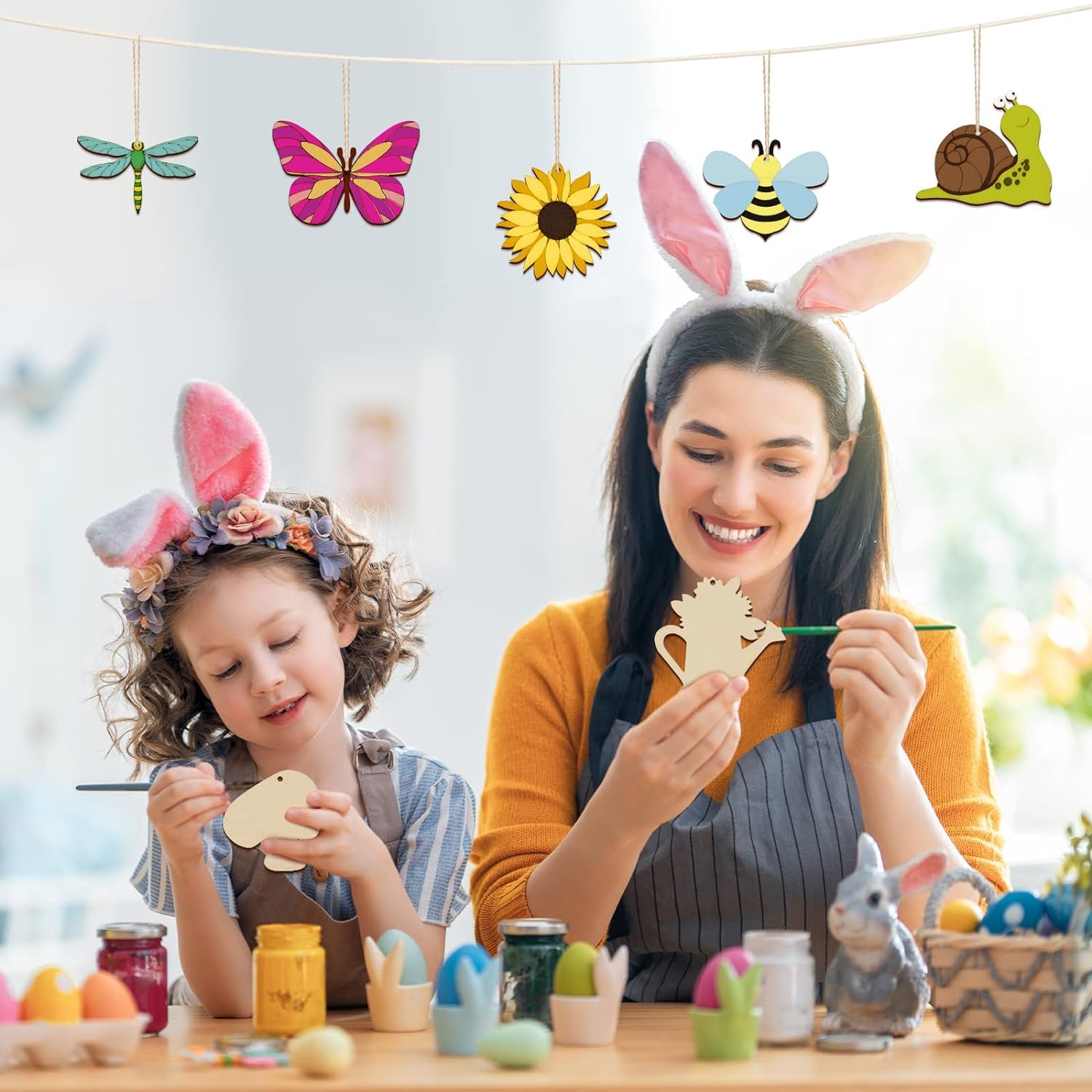 Woman and child wearing bunny ears, painting Easter eggs in a bright room.