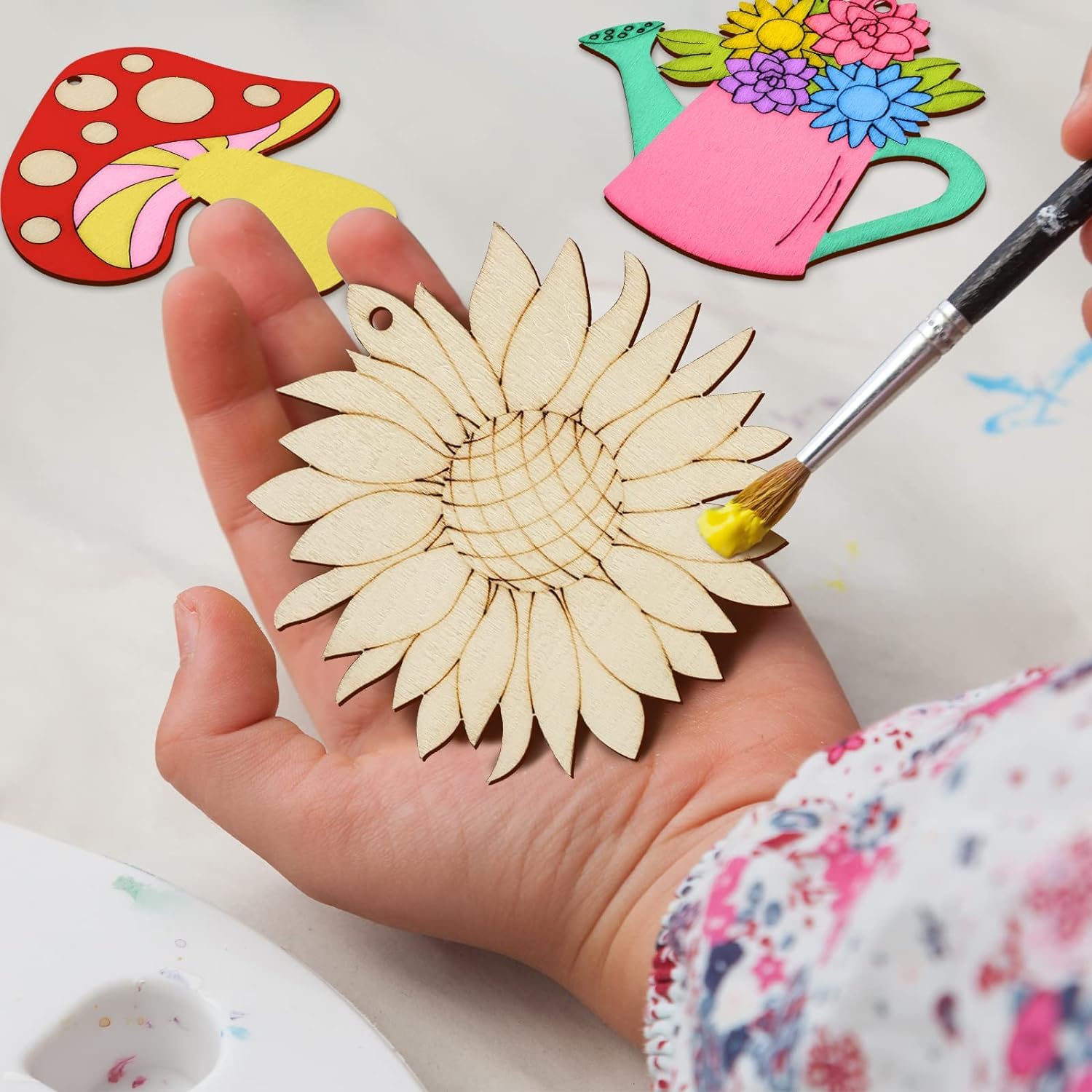 Hand holding a wooden sunflower ornament with paintbrush and colorful craft items in the background